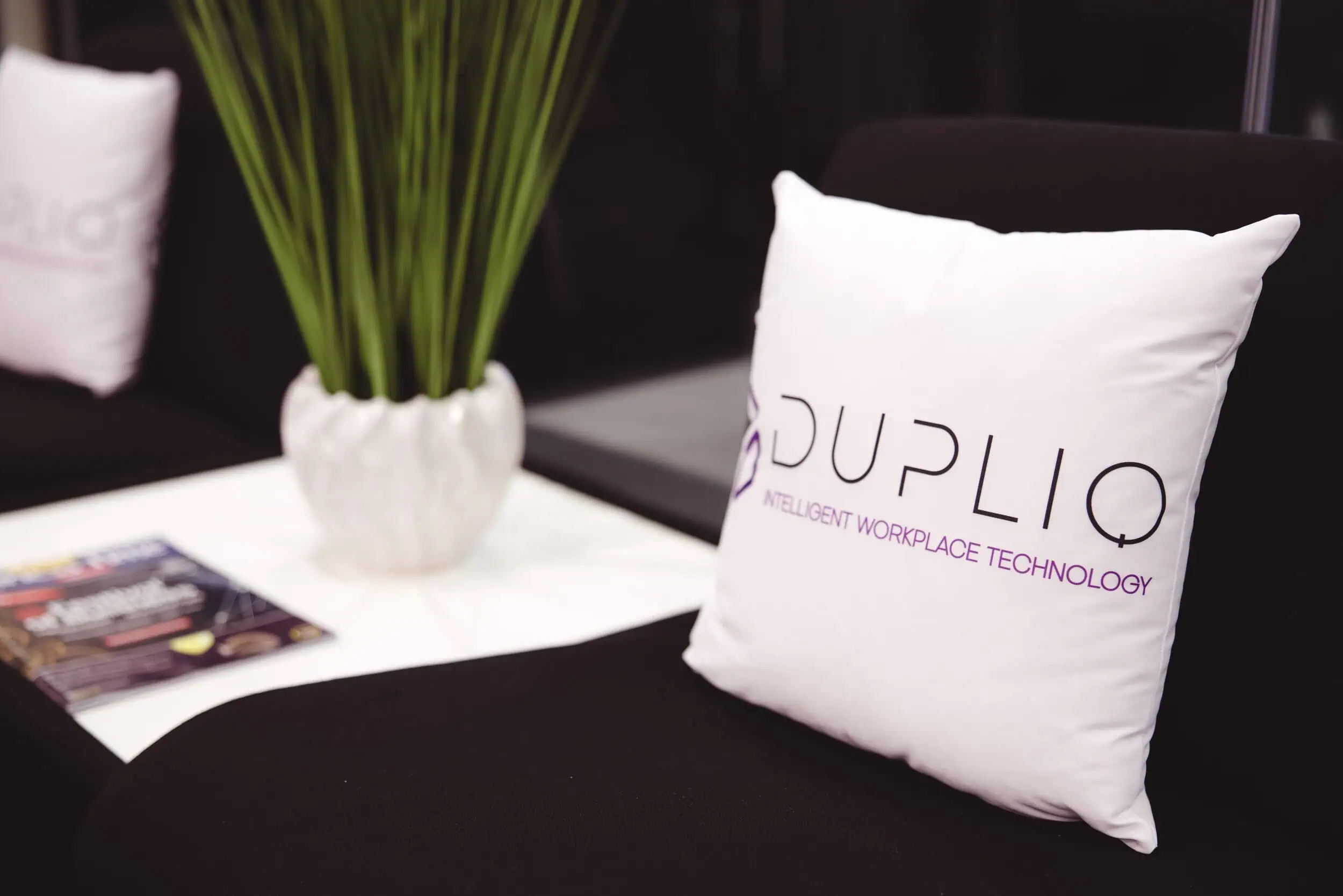 DUPLIQ branded cushion on a dark armchair beside a plant in an office lounge area