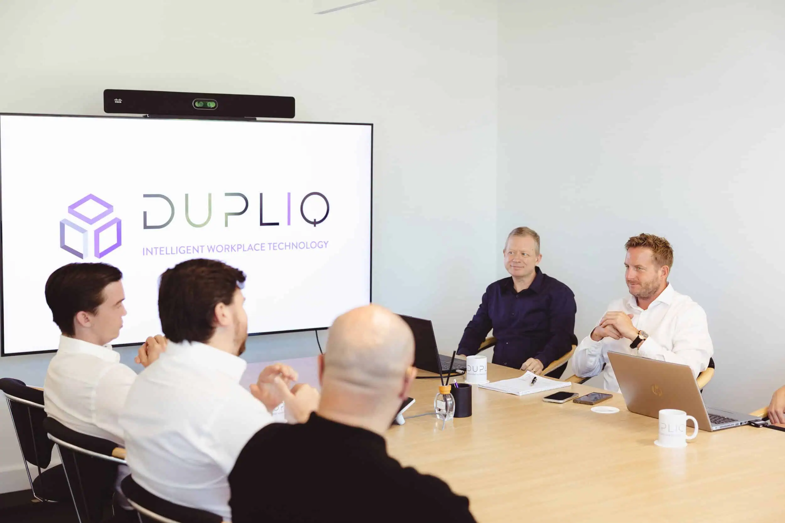 DUPLIQ leadership team meeting in a boardroom with laptops and the DUPLIQ logo displayed